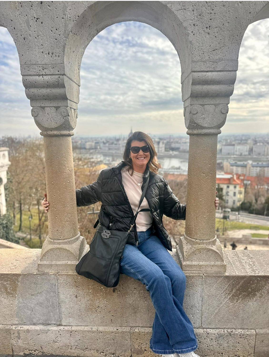 Anna-Swenson-209 A woman in sunglasses and a black jacket sits on a stone structure framed by two arches, with a cityscape and river in the background under a cloudy sky.