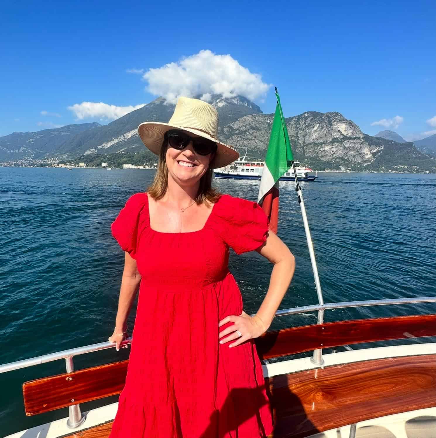 Anna-Swenson-210 Woman in a red dress and sunhat on a boat with an Italian flag, with mountains and a lake visible in the background under a clear blue sky.