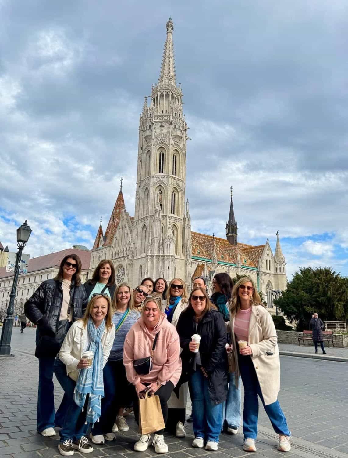 Anna-Swenson-211 A group of people pose and smile in front of a tall, ornate church with a pointed spire under a cloudy sky.