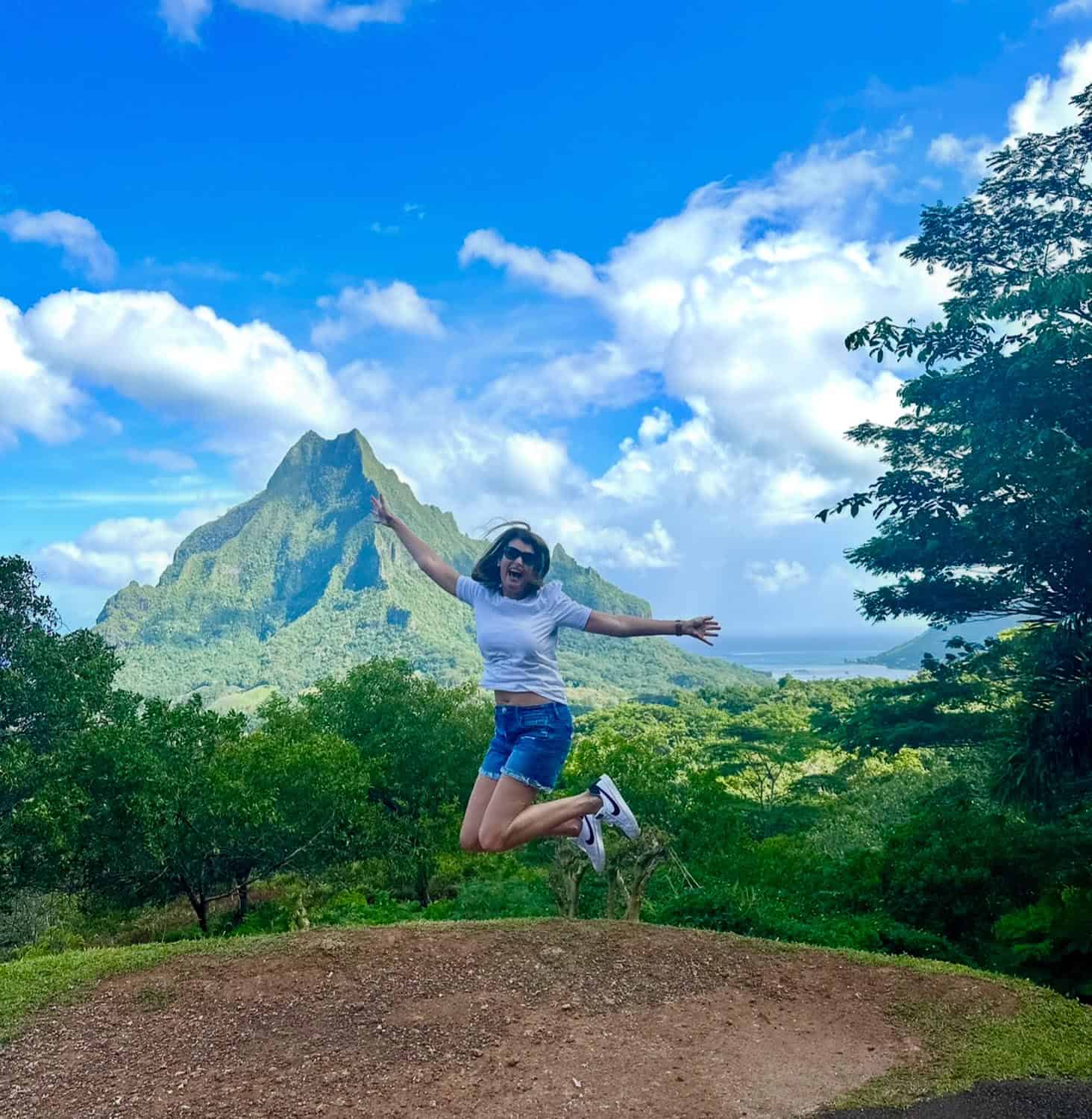 Anna-Swenson-212 A person wearing a white shirt and shorts jumps in the air with arms outstretched in front of a lush mountain landscape under a blue sky with clouds.