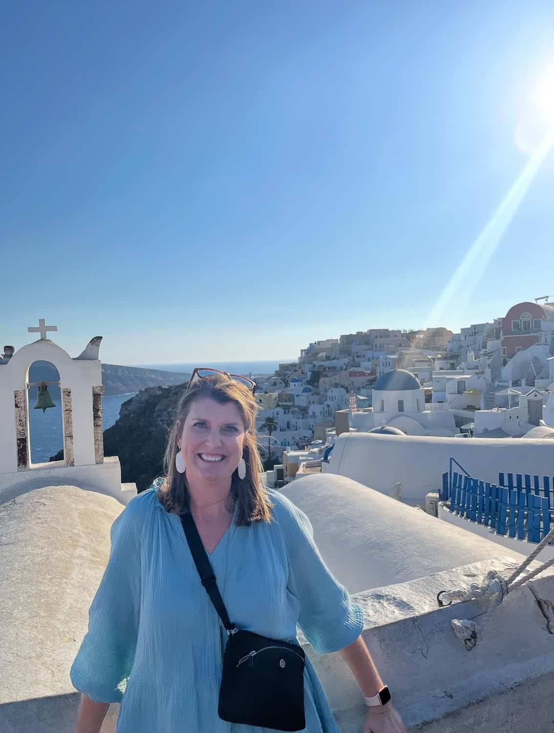 Anna-Swenson-213 Woman in a light blue dress stands on a white rooftop in Santorini, Greece, with white buildings and blue sea in the background under a clear, sunny sky.