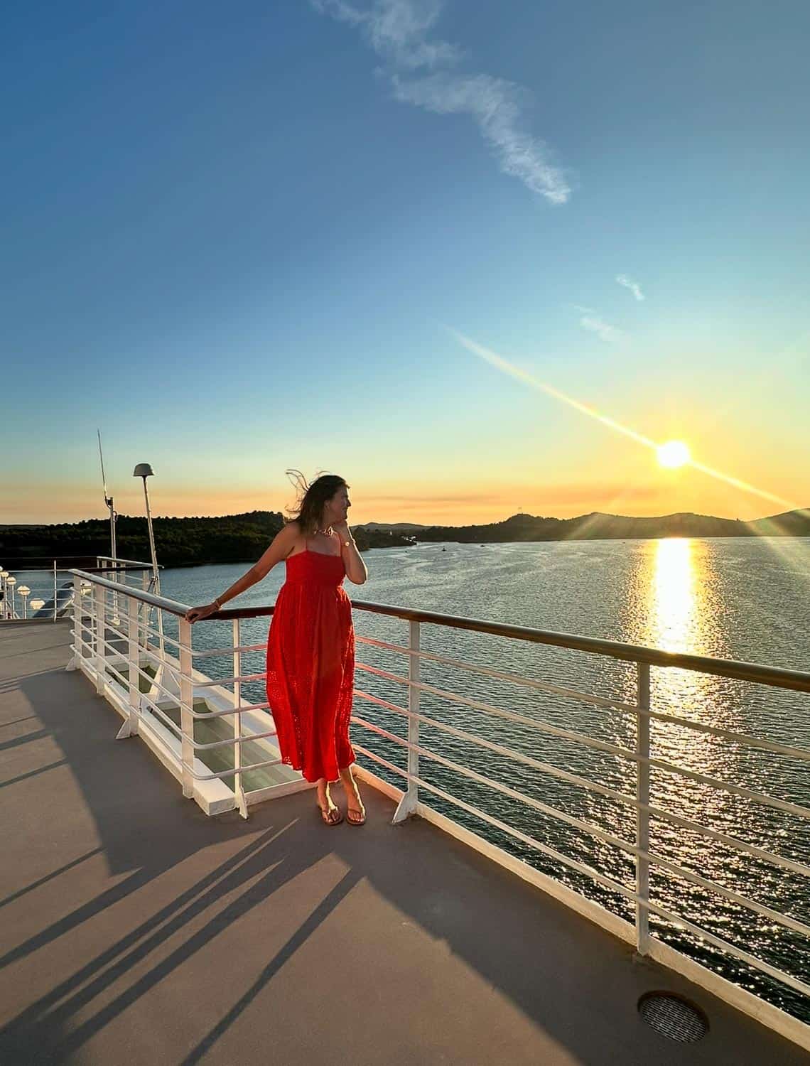 Anna-Swenson-214 A woman in a red dress stands on a cruise ship deck overlooking the water at sunset, the sun low on the horizon and calm surroundings.