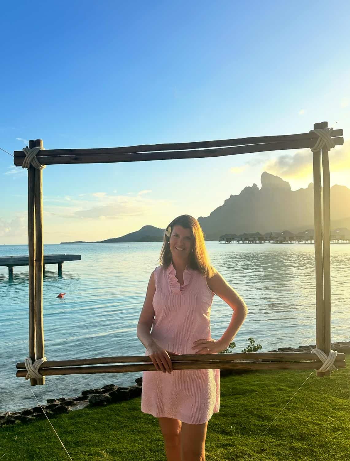 Anna-Swenson-216 Woman in pink sleeveless dress stands behind a large wooden frame by the waterfront, with mountains and overwater bungalows in the sunset background.