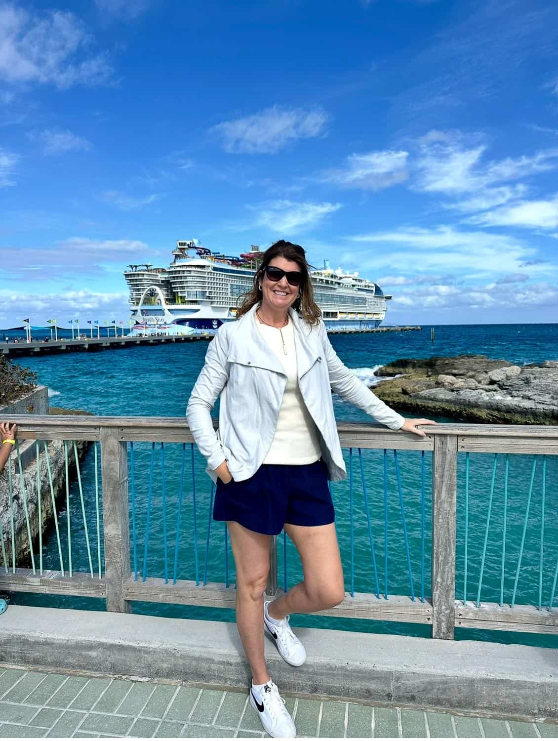 Anna-Swenson-217 Woman standing by a wooden railing near the ocean with a large cruise ship in the background under a blue sky with clouds.
