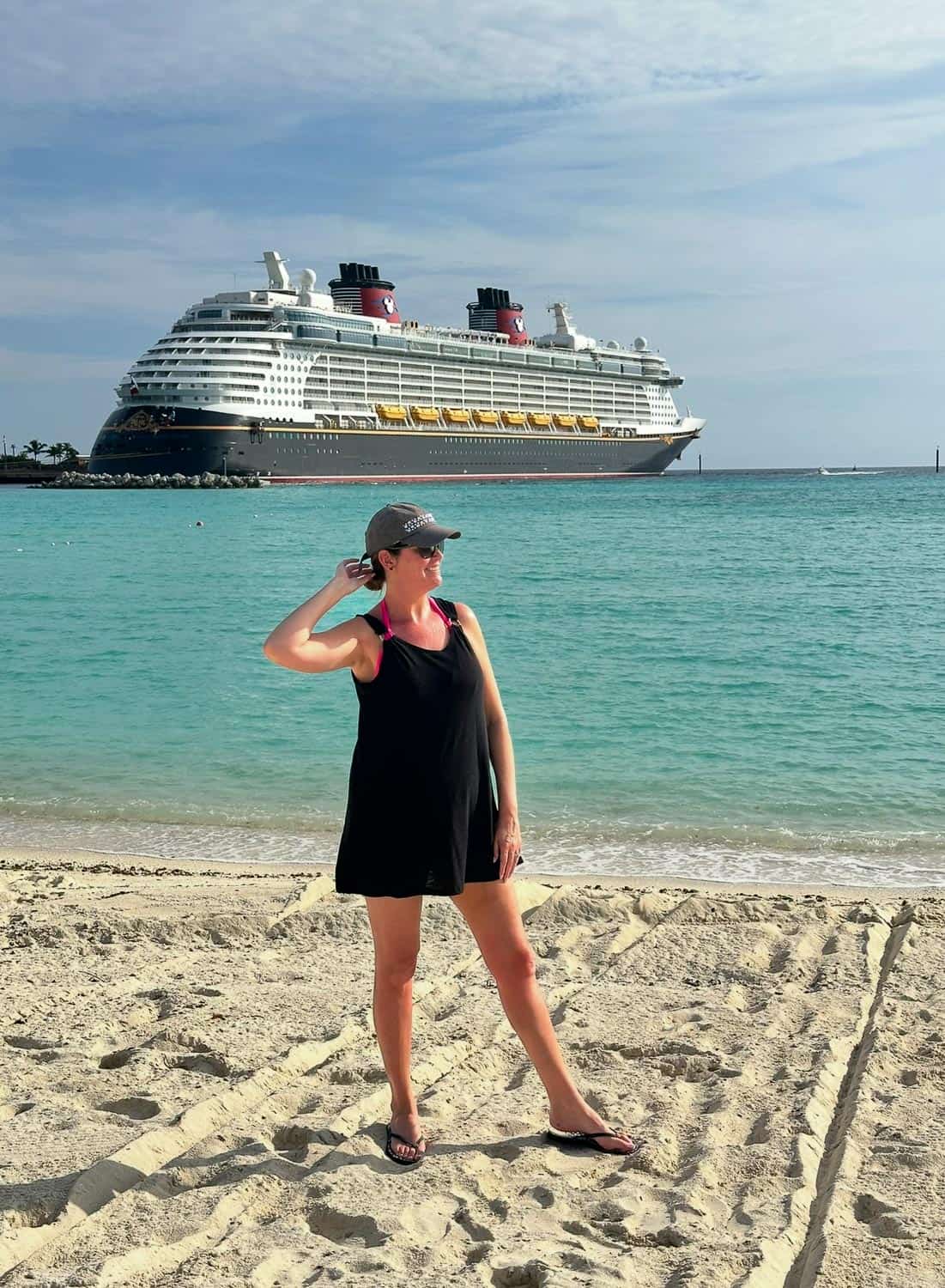 Anna-Swenson-218 A woman in a black dress and gray cap stands on a sandy beach with a large cruise ship in the turquoise water behind her.