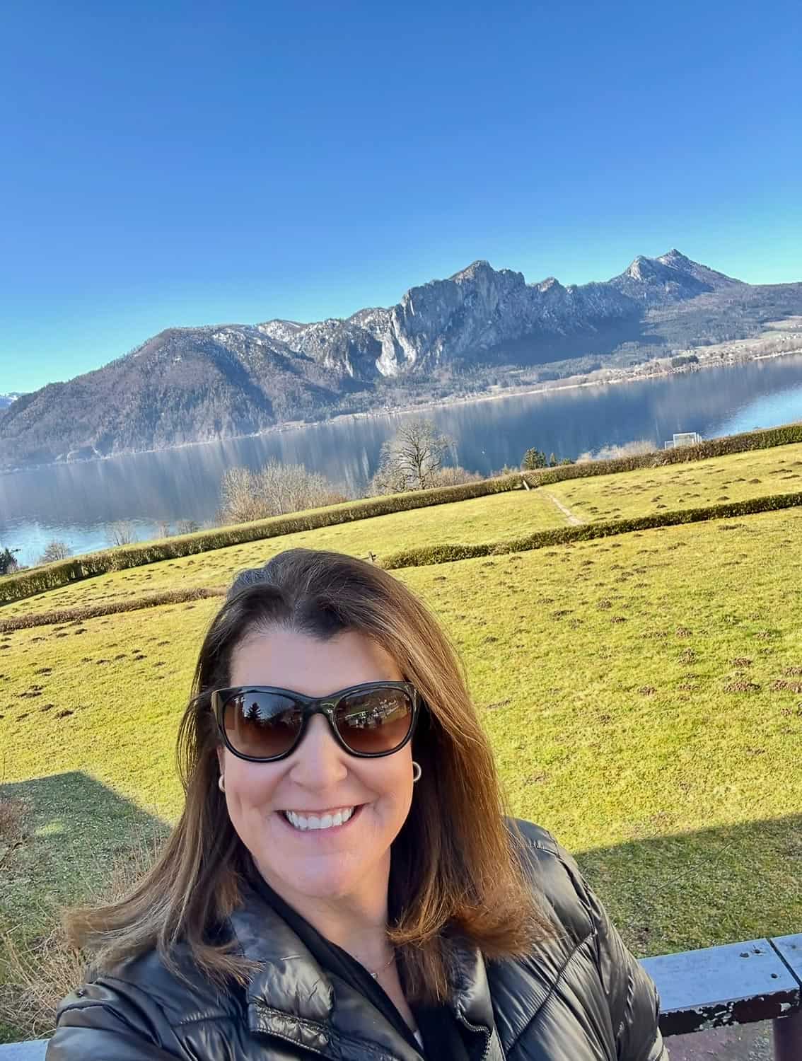 Anna-Swenson-219 A woman wearing sunglasses and a black jacket smiles for a selfie outdoors with a lake, grassy field, and mountains in the background.