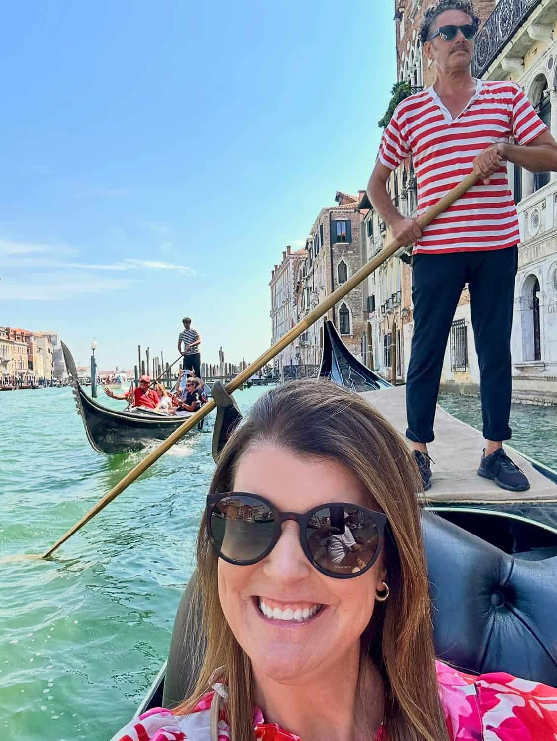Anna-Swenson-220 A woman takes a selfie on a gondola in Venice, Italy, with a gondolier standing behind her and other gondolas on the canal.