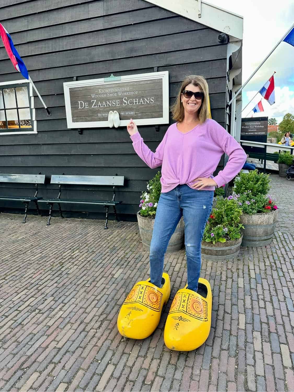 Anna-Swenson-222 A woman in a purple top and jeans poses in oversized yellow wooden clogs in front of a building with a sign reading De Zaanse Schans in the Netherlands.