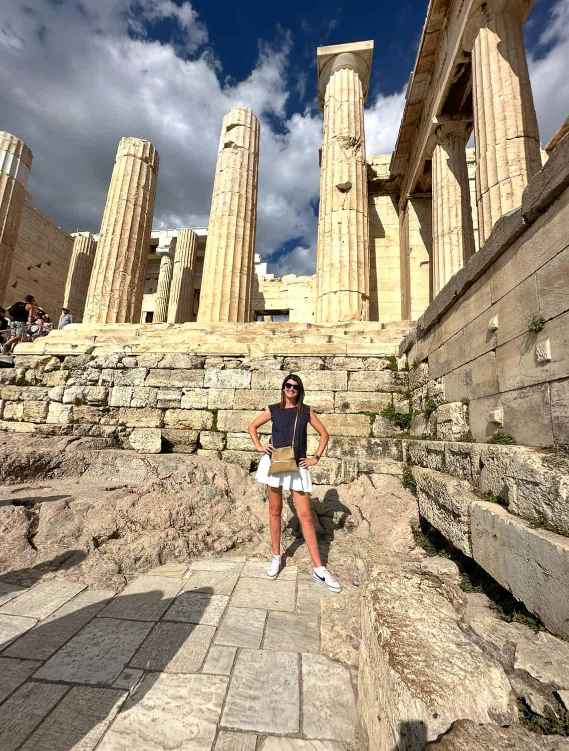 Anna-Swenson-223 A woman stands in front of ancient stone columns and ruins in Italy under a partly cloudy sky.
