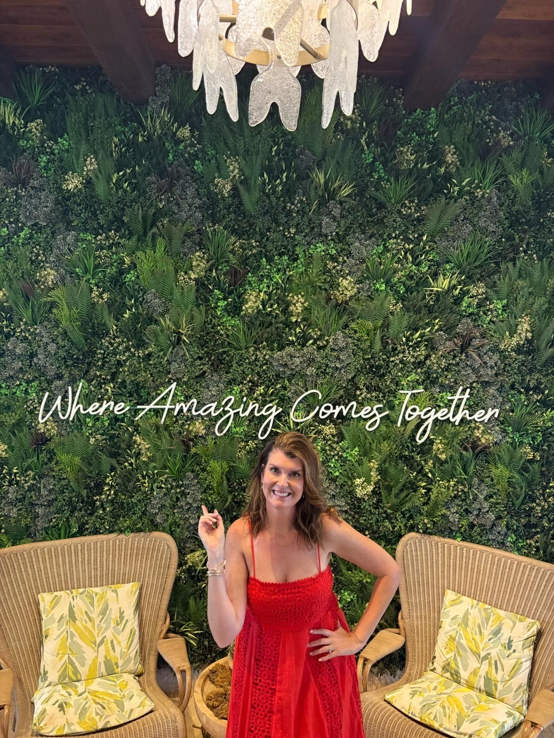 Anna-Swenson-224 A woman in a red dress in front of a green plant wall with the words Where Amazing Comes Together, flanked by two wicker chairs with leaf-patterned cushions.