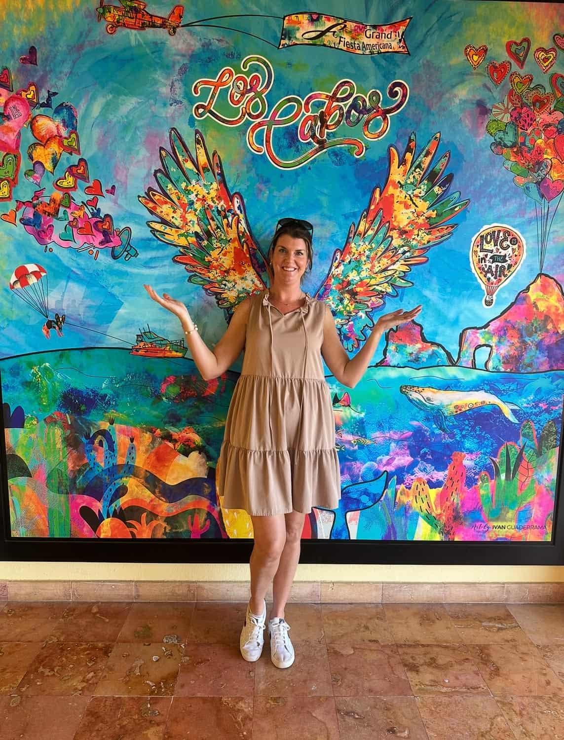 Anna-Swenson-225 A woman in a beige dress stands in front of a colorful Los Cabos mural with painted wings that appear to be attached to her back.