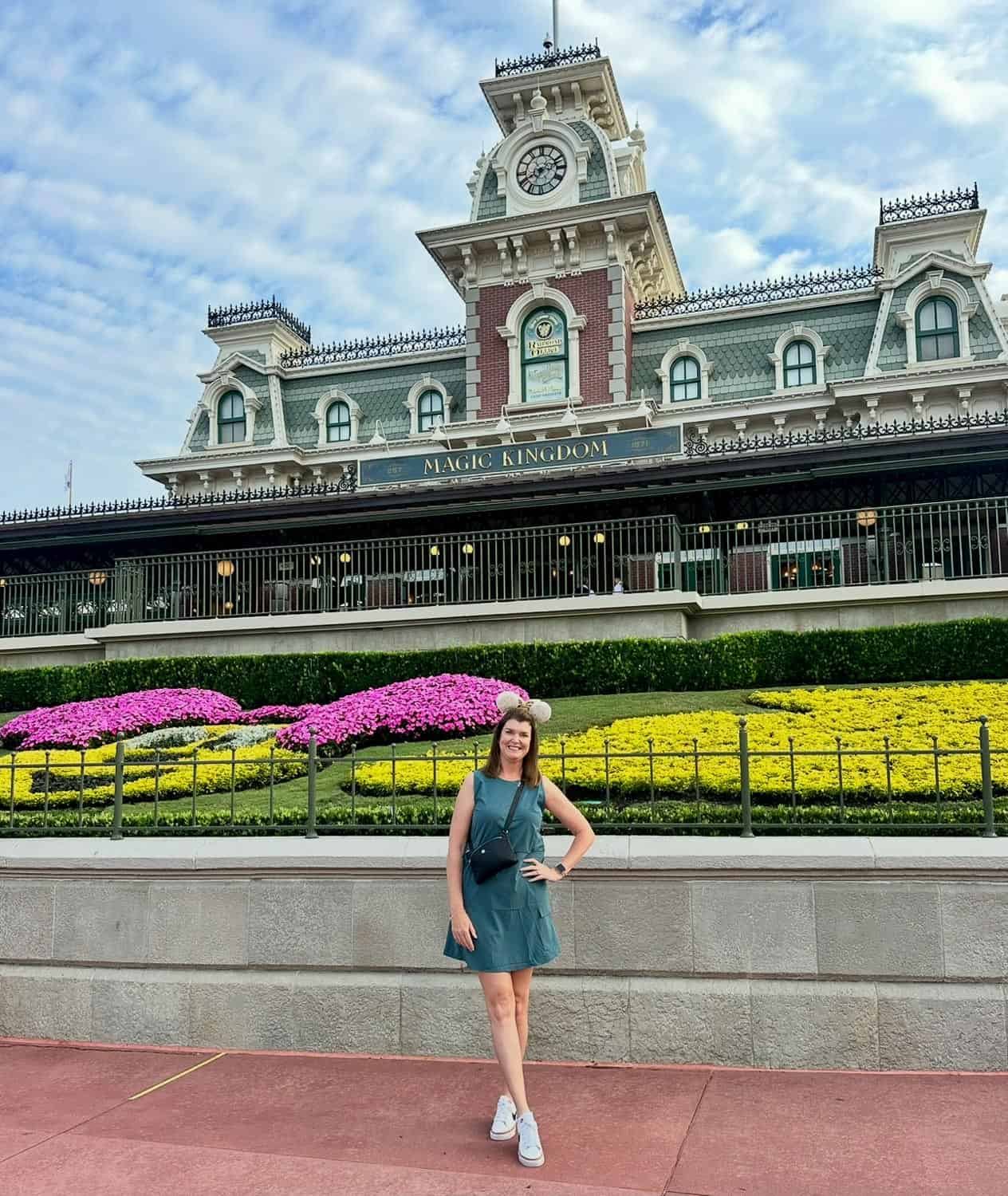 Anna-Swenson-227 A woman in a green dress stands smiling in front of the Magic Kingdom entrance at Walt Disney World, with the train station and flower beds in the background.