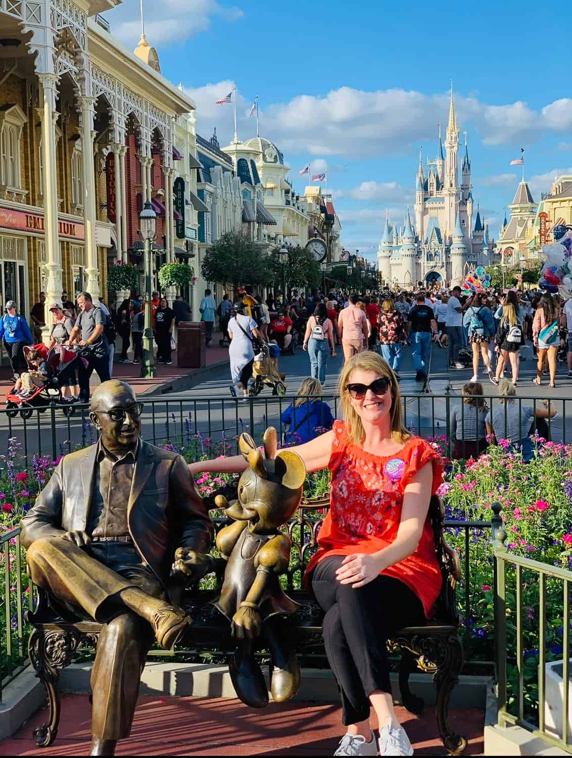 Anna-Swenson-228 A woman sits on a bench next to statues of Walt Disney and Minnie Mouse on Main Street, with Cinderella Castle and crowds in the background.