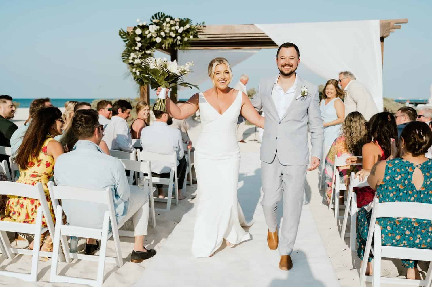 A bride and groom walk down the aisle outdoors, smiling, while guests seated on white chairs watch and celebrate.