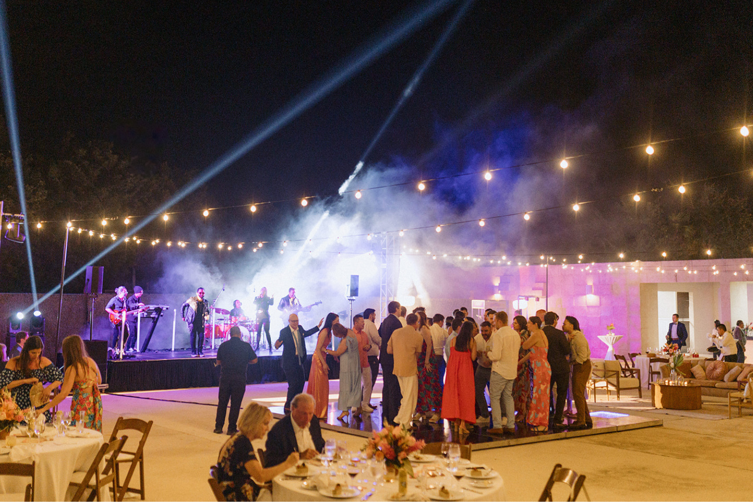 A bride and groom stand on a rooftop, embracing and kissing. The bride’s veil flows in the wind, and a scenic view is visible in the background.