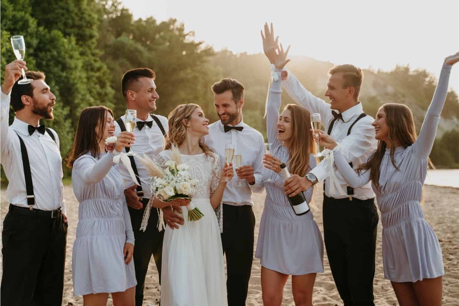 Couple kisses under a floral arch at an outdoor beach destination wedding, surrounded by cheering guests, palm trees, and white flowers.